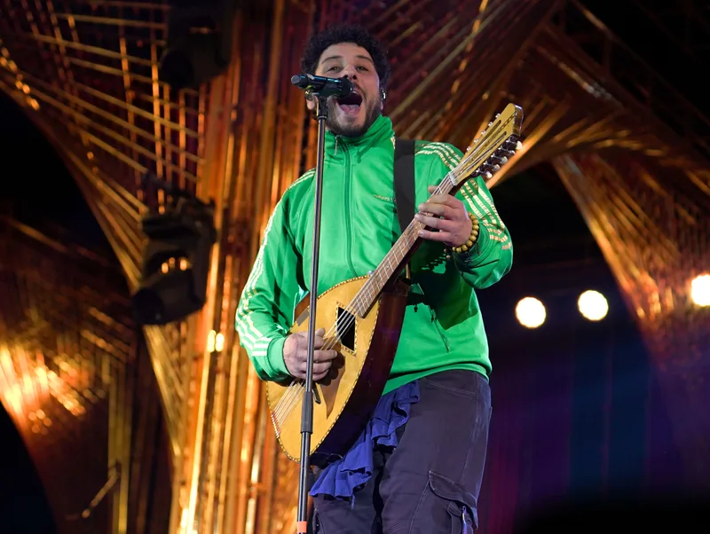 Singer in a green jacket playing a string instrument beneath a wooden canopy